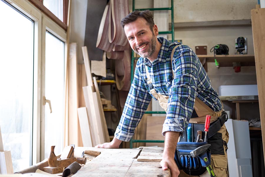 Business Insurance - Young Contractor Smiles Over His Workbench With Tools Surrounding Him, Wearing Blue Flannel and a Tool Belt