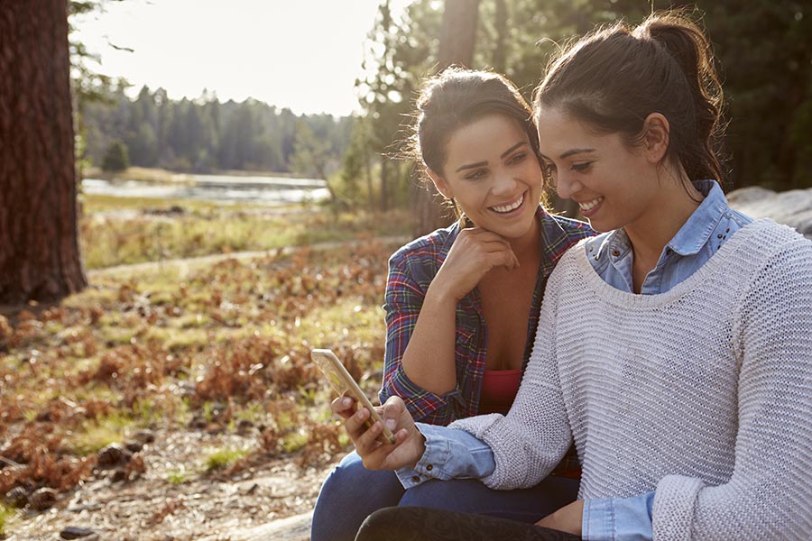 Contact Us - Couple in Fall Clothing Smiles and Uses Phone Seated by a Lake With Pine Trees Surrounding Them